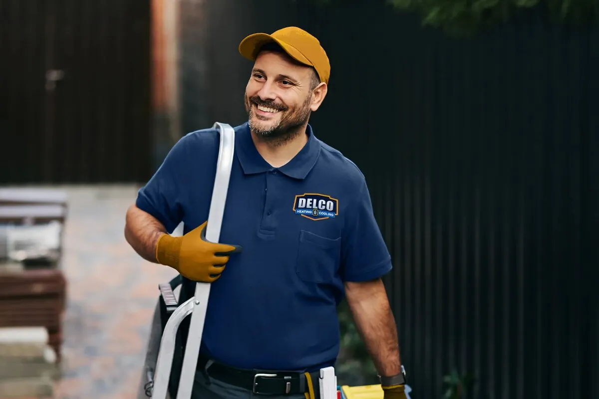 A uniformed HVAC technician carrying equipment beside a house